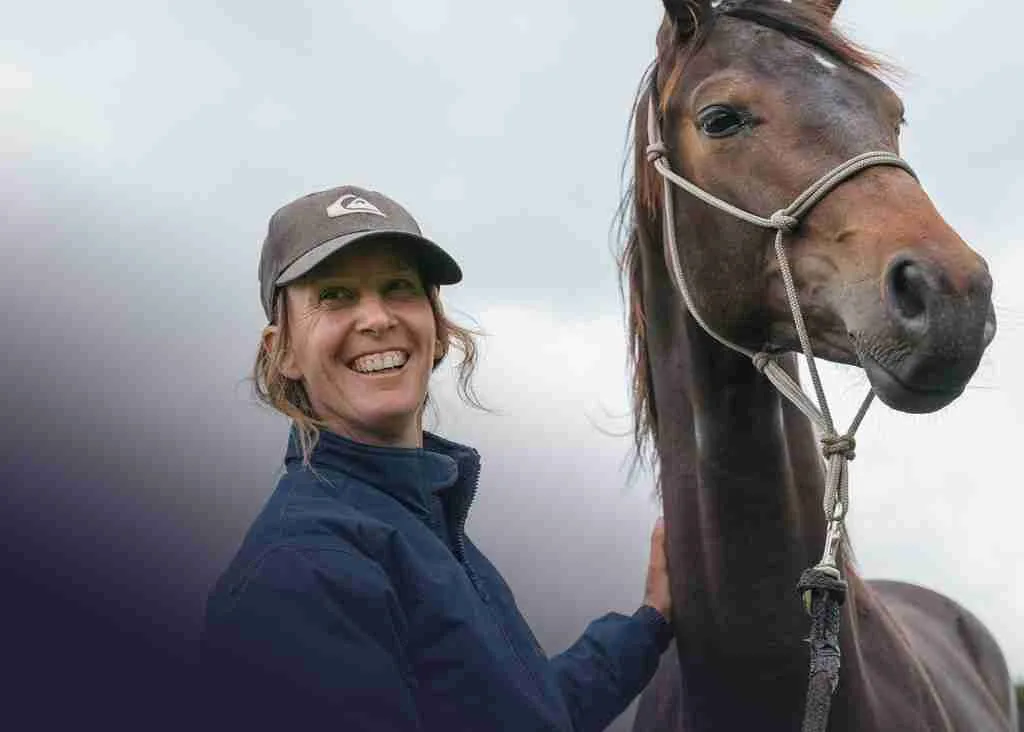 A trainer in a cap smiling beside a dark bay horse held in a rope halter